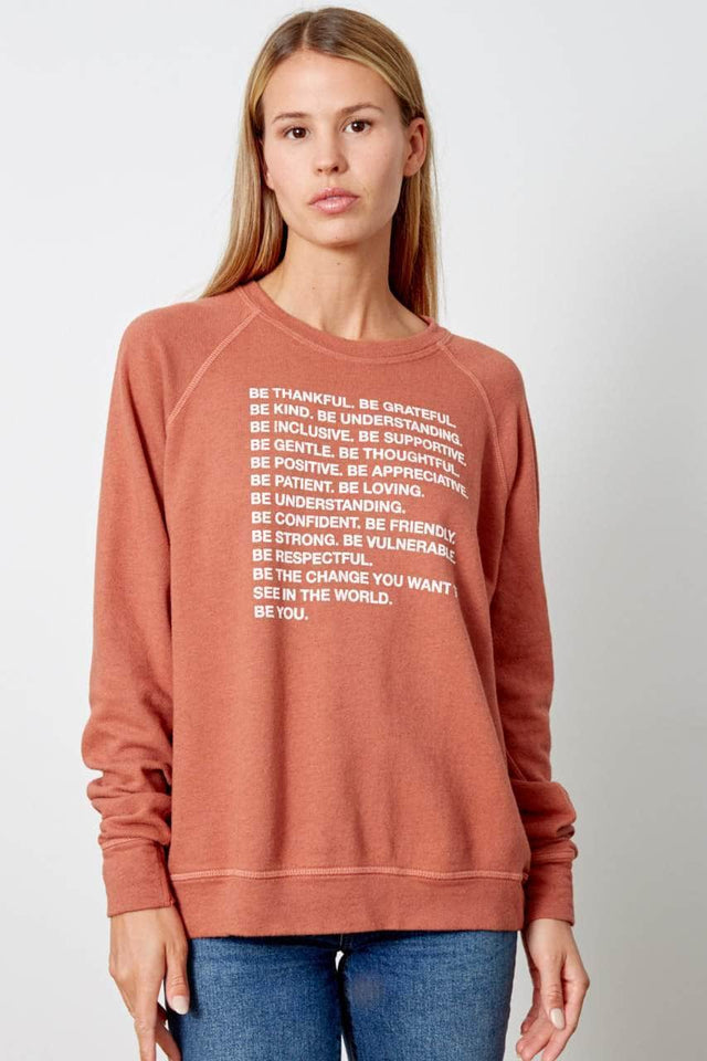 burnt orange sweatshirt with raglan sleeves. Fits loose and hits just past the hip. Graphic printed in white on front reads "Be Thankful. Be Grateful. Be Kind. Be Understanding. Be Inclusive. Be Supportive. Be Gentle. Be Thoughtful. Be Positive. Be Appreciative. Be Patient. Be Loving. Be Understanding. Be Confident. Be Friendly. Be Strong. Be Vulnerable. Be Respectful. Be the change you want to see in the world. be YOU. 