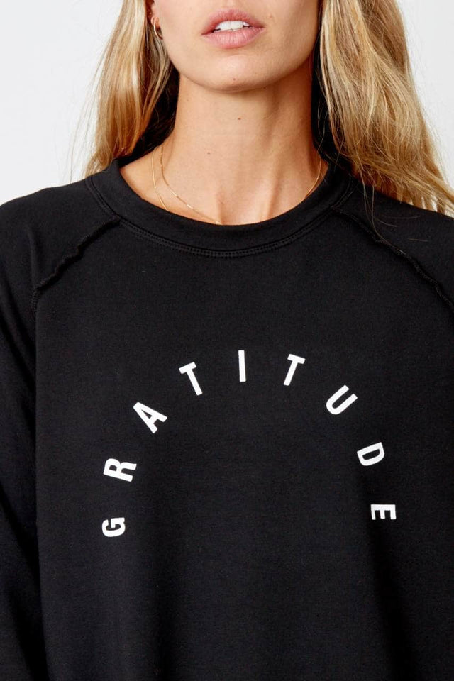 Model is wearing the Vita, a black crewneck sweatshirt with raglan sleeves that hits just past the hips. The word Gratitude is printed in an arc shape across the chest in white ink.